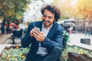 Mann in blauen Blazer sitzt auf einer Bank und tippt lachend in sein Handy